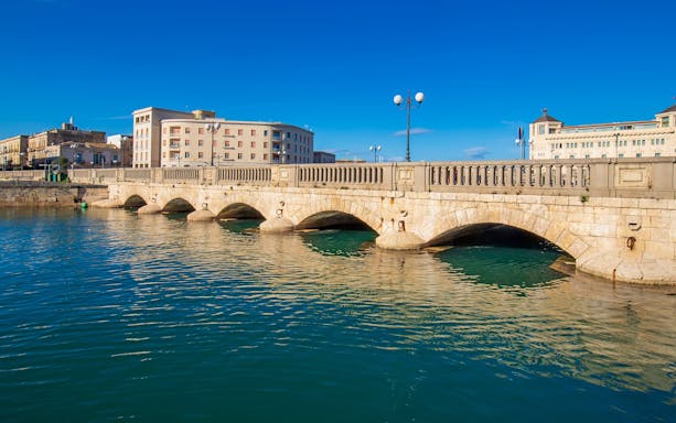 Umberto I Bridge over water in Syracuse, Sicily, Italy with buildings in the background.