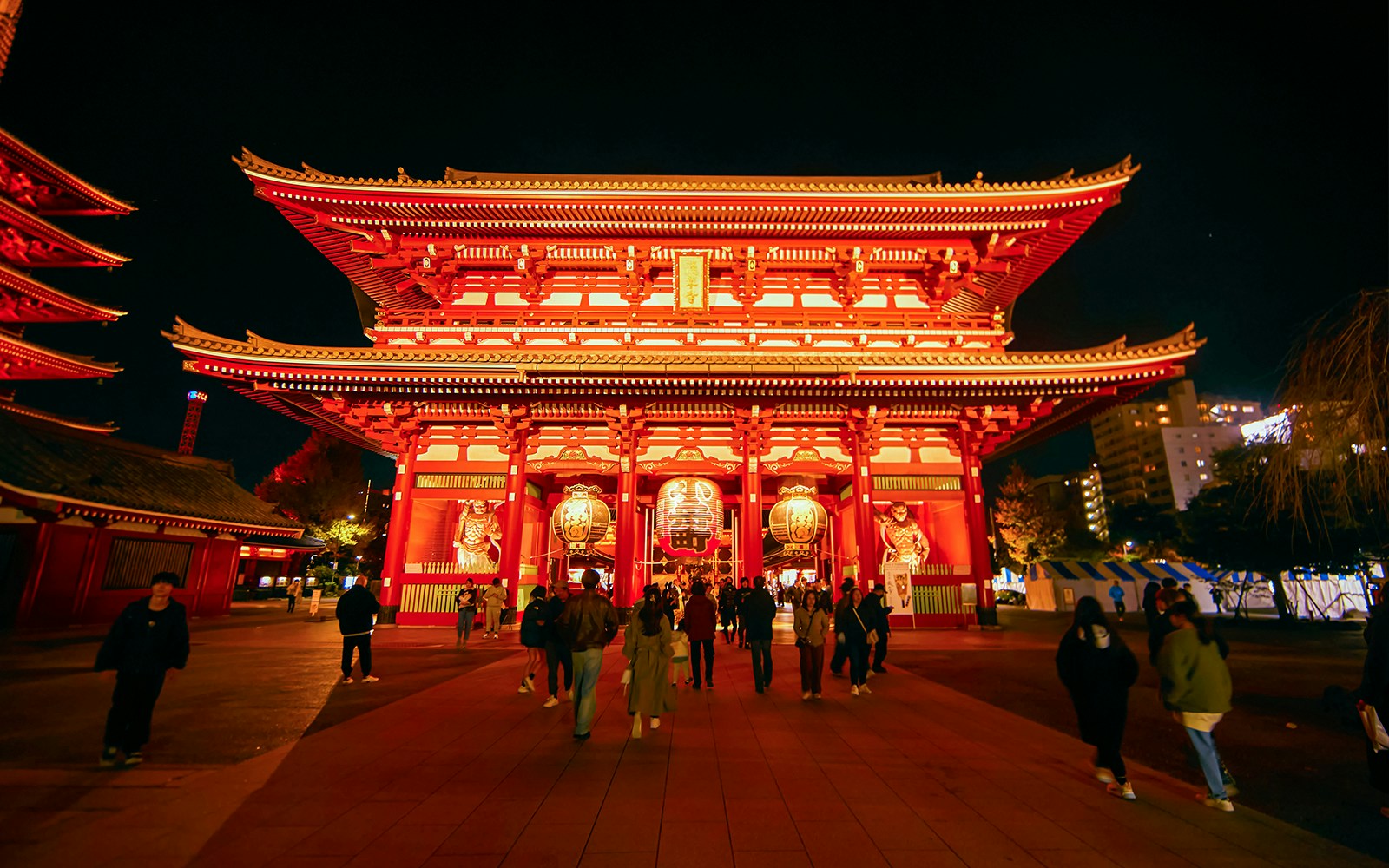 Sensoji Temple illuminated at night in Asakusa, Tokyo, with visitors walking towards the entrance.