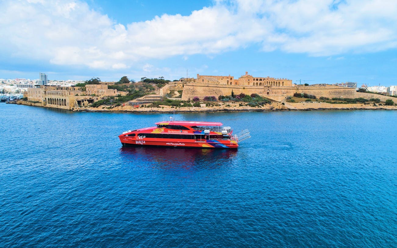 Red tour boat cruising near historic fortifications in Malta's harbor.