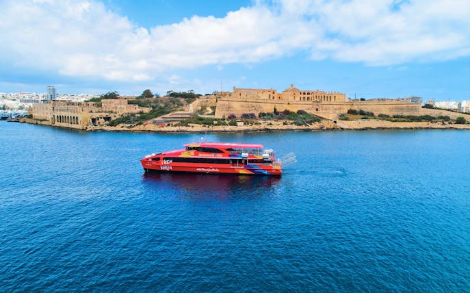 Red tour boat cruising near historic fortifications in Malta's harbor.