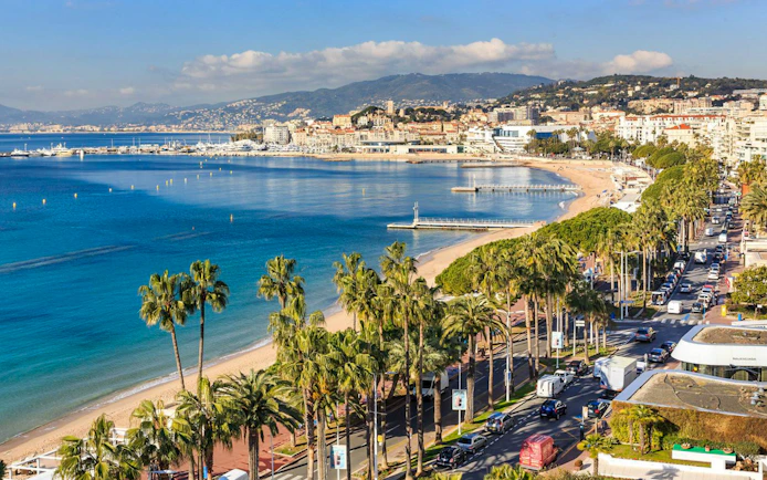 Cannes coastline with palm-lined promenade and beach, view from the half-day tour from Nice.