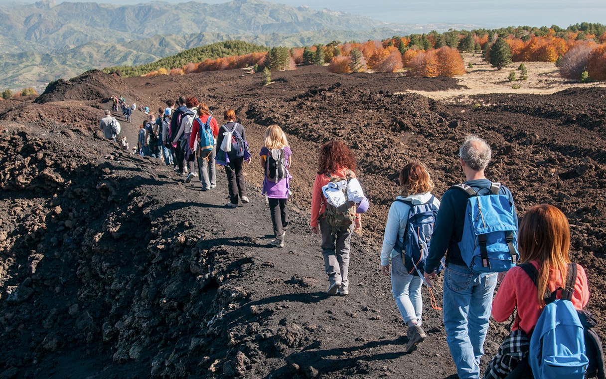 Hikers walking on a volcanic trail towards Mount Etna, Sicily, with autumn foliage in the background.