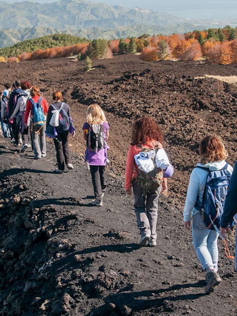 Hikers walking on a volcanic trail towards Mount Etna, Sicily, with autumn foliage in the background.