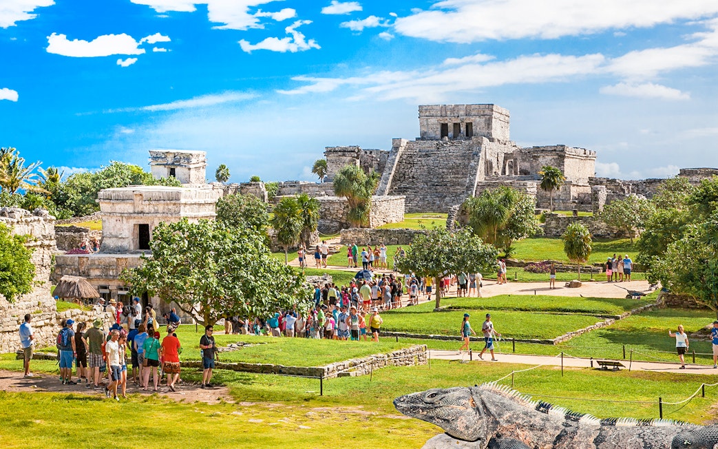 Iguana on Tulum ruins with tourists exploring ancient site in Mexico.