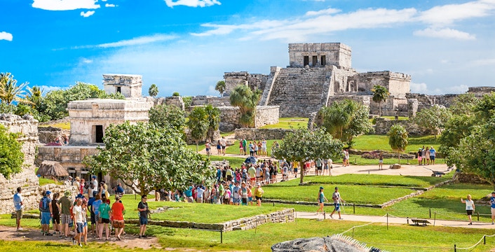 Iguana on Tulum ruins with tourists exploring ancient site in Mexico.