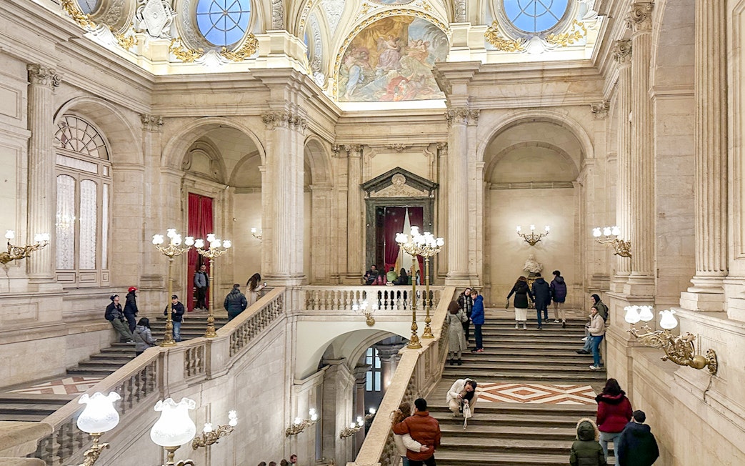 Main staircase of the Royal Palace of Madrid with visitors exploring the ornate architecture.
