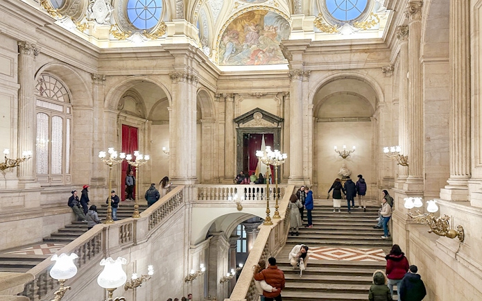 Main staircase of the Royal Palace of Madrid with visitors exploring the ornate architecture.
