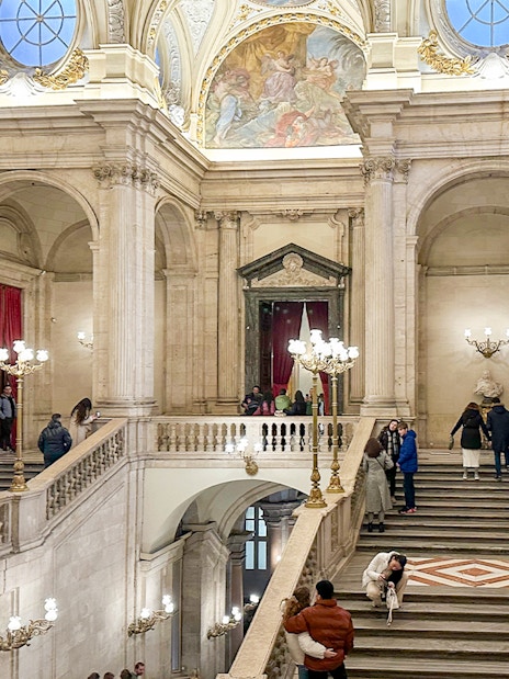 Main staircase of the Royal Palace of Madrid with visitors exploring the ornate architecture.