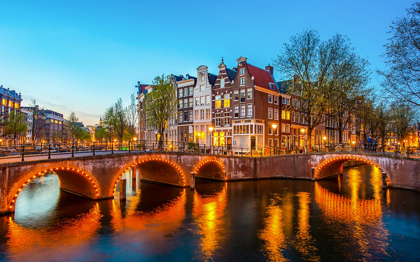 Canal bridge illuminated at night in Amsterdam, Netherlands, with traditional houses lining the water.