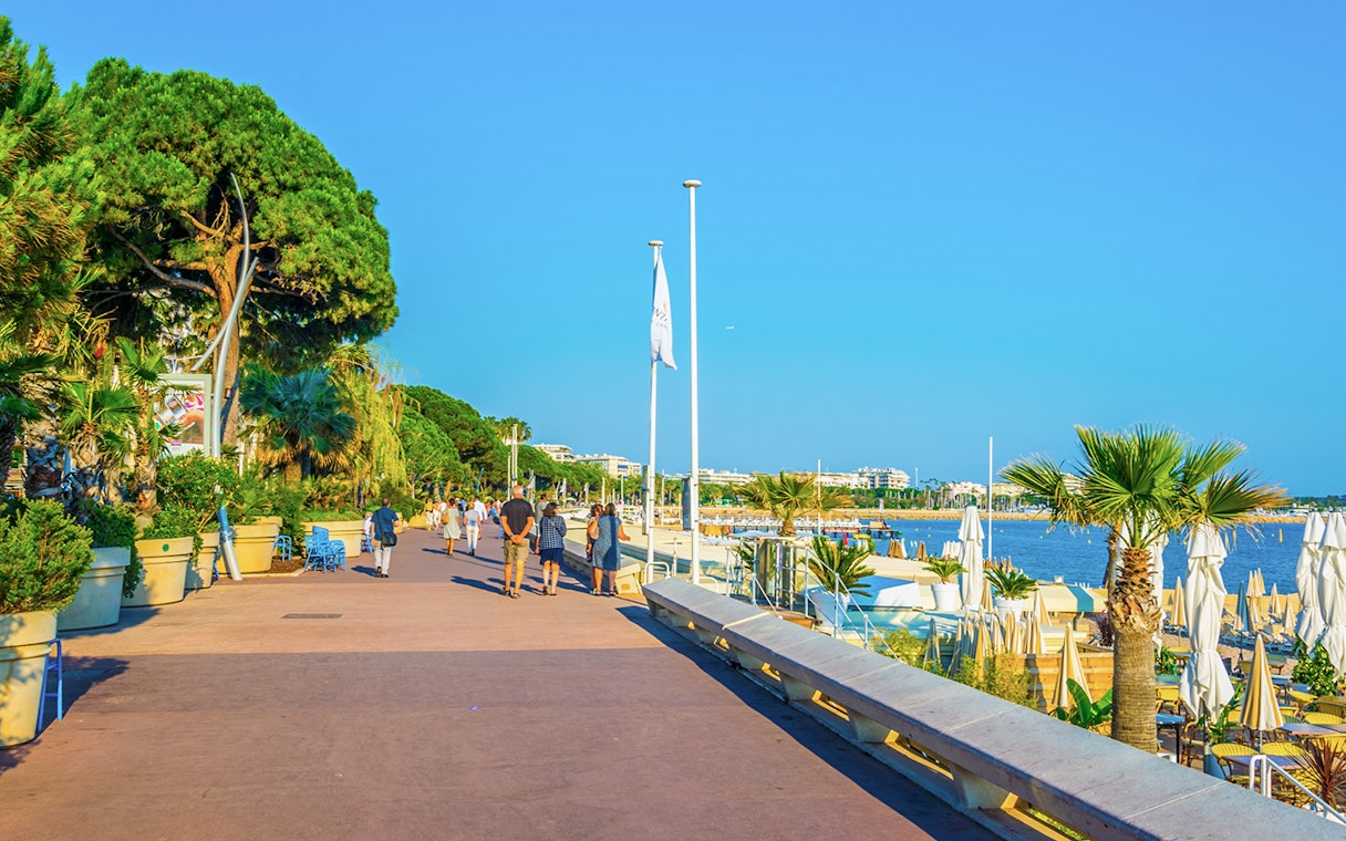 People walking along boulevard de la Croisette in Cannes, near the beach and palm trees.