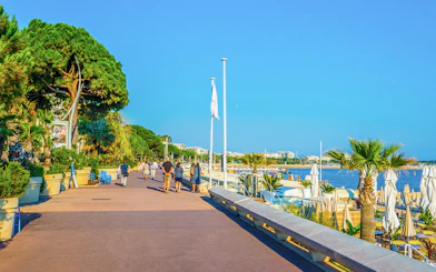 People walking along boulevard de la Croisette in Cannes, near the beach and palm trees.
