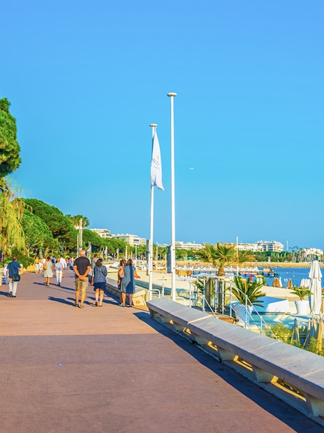 People walking along boulevard de la Croisette in Cannes, near the beach and palm trees.