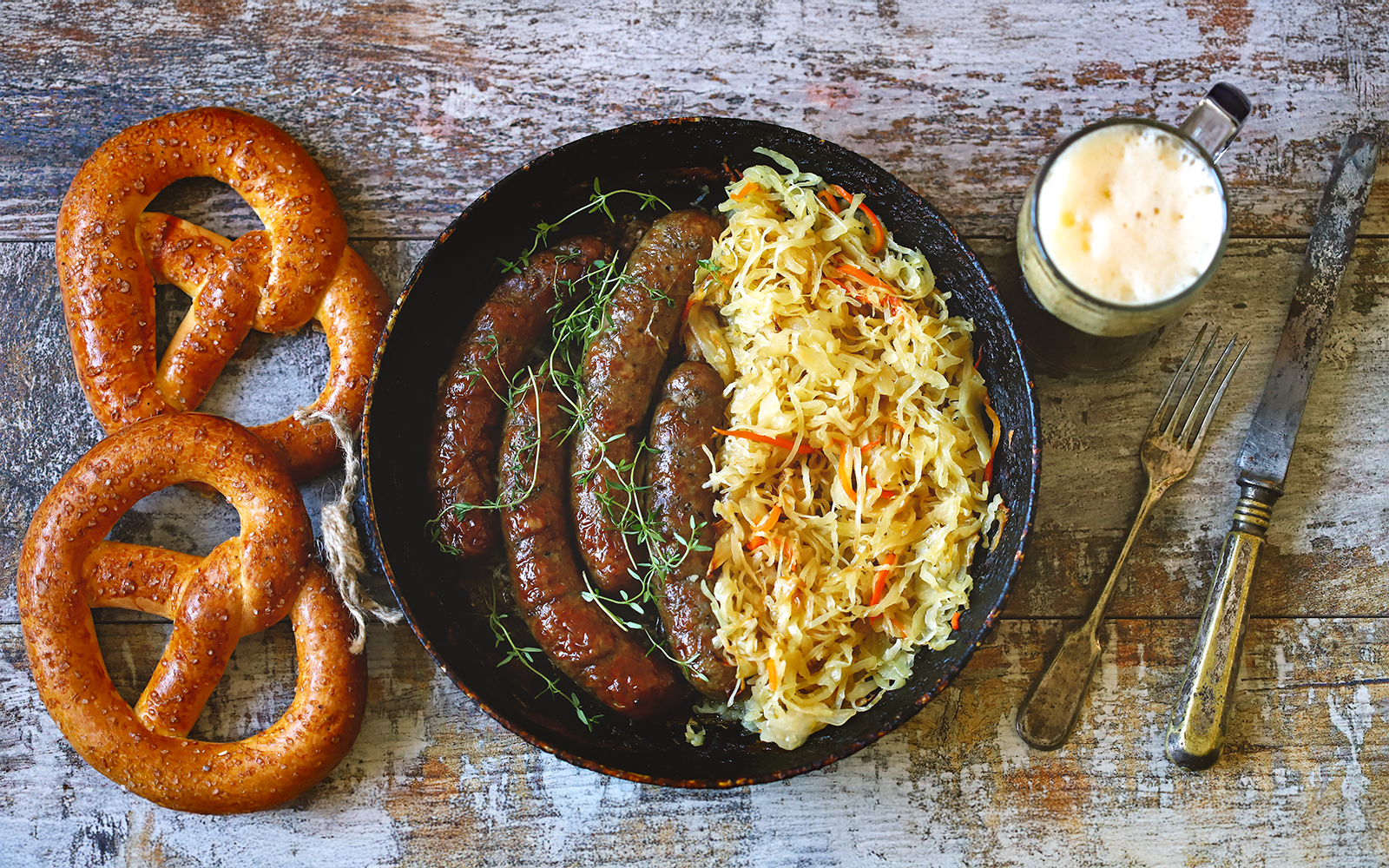 Bratwurst with sauerkraut, pretzels, and a mug of beer on a rustic table.