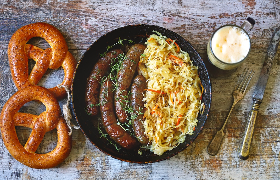 Bratwurst with sauerkraut, pretzels, and a mug of beer on a rustic table.