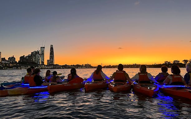 Kayakers with LED lights paddle on Sydney Harbour at sunset, city skyline silhouette.