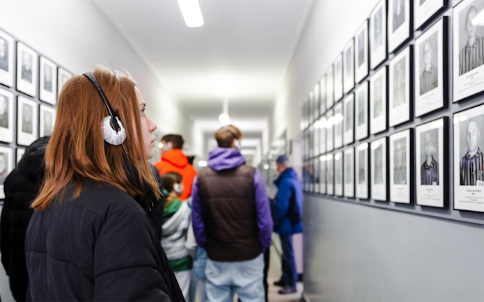 Visitor with audio guide at Auschwitz-Birkenau Memorial, viewing prisoner photos in a hallway.