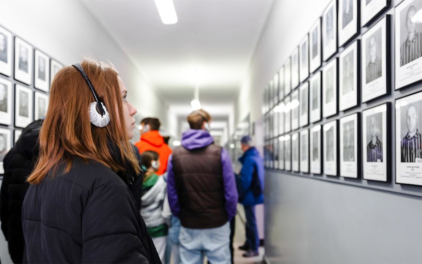 Visitor with audio guide at Auschwitz-Birkenau Memorial, viewing prisoner photos in a hallway.