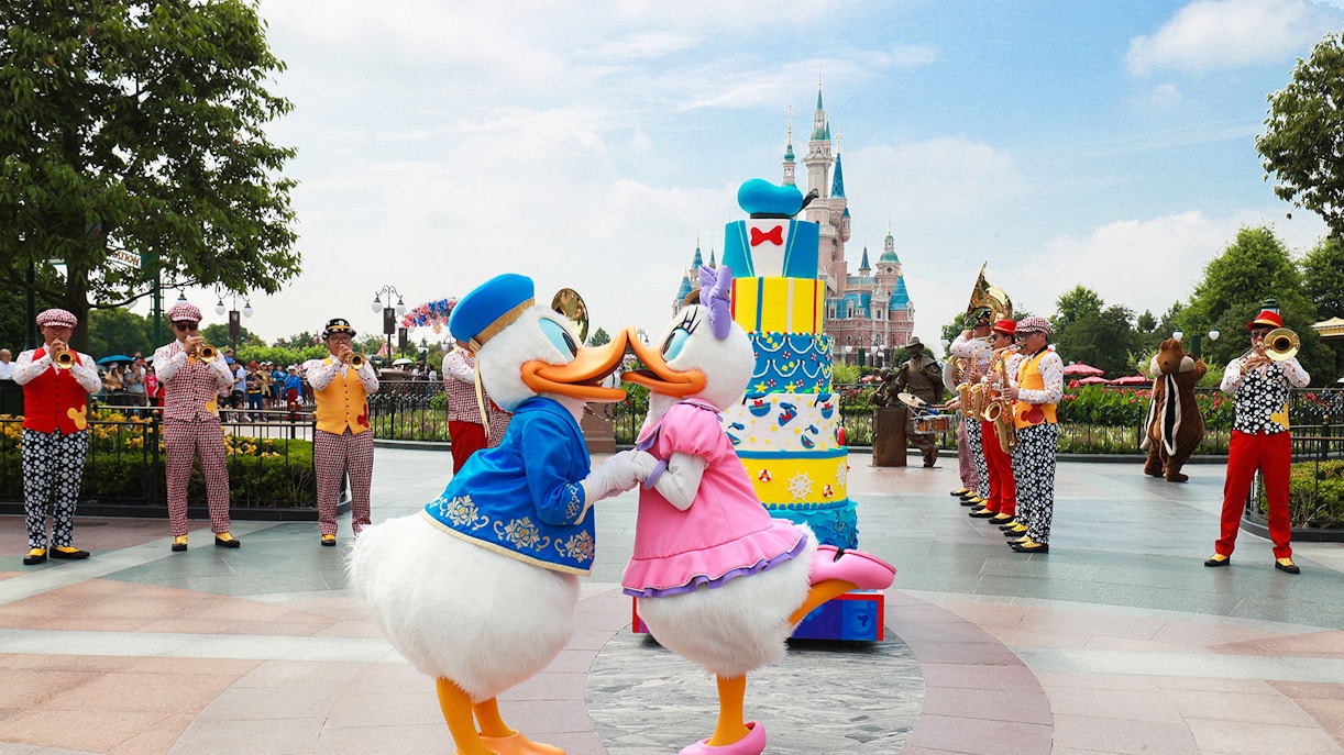 Donald and Daisy Duck dancing in front of a cake at Shanghai Disneyland with a band playing nearby.