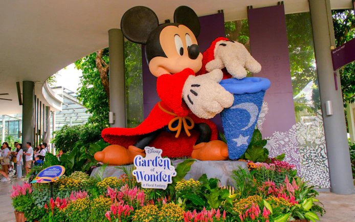 Mickey Mouse statue in red robe at Disney Garden of Wonder, surrounded by colorful flowers.