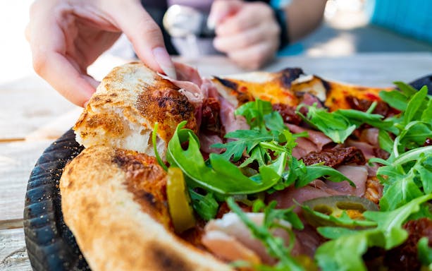 Hand picking slice of pizza with arugula and prosciutto in Naples street food tour.