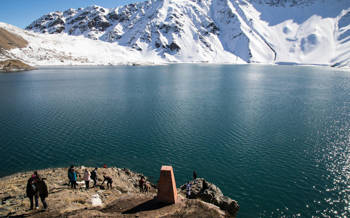 Tourists exploring the shoreline of Embalse El Yeso with snow-capped mountains in the background.