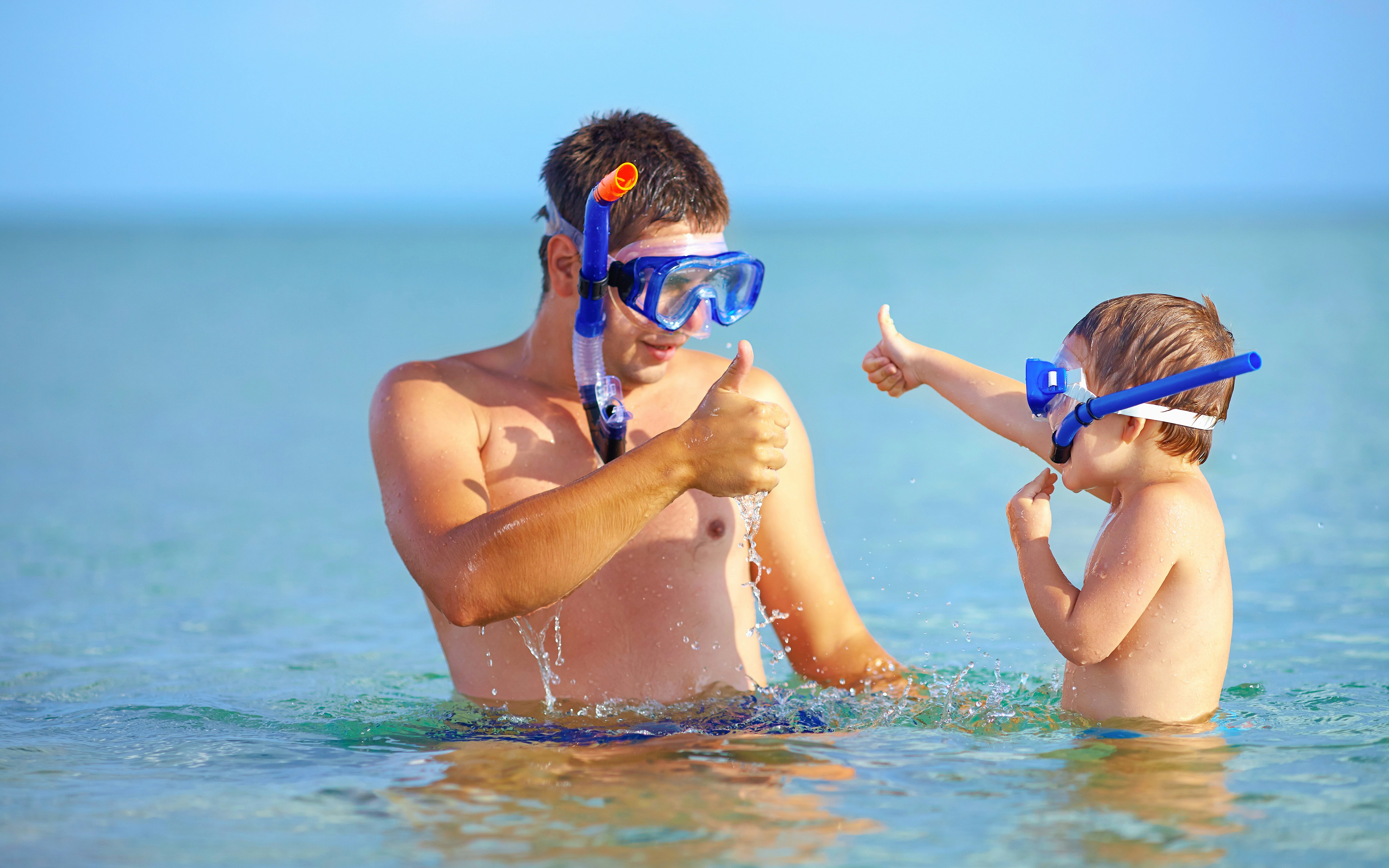 Father and son giving thumbs up while snorkeling in clear ocean water.