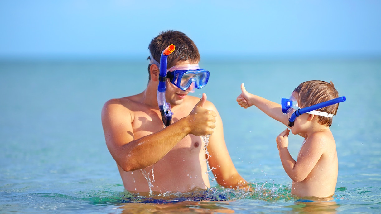 Father and son giving thumbs up while snorkeling in clear ocean water.