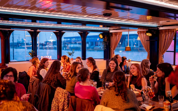 Group enjoying pizza on a river cruise with cityscape views through large windows.