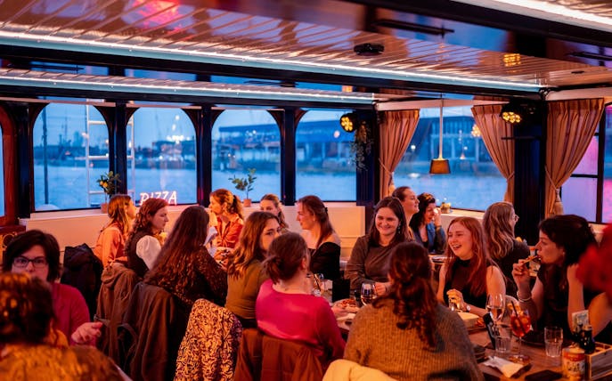 Group enjoying pizza on a river cruise with cityscape views through large windows.