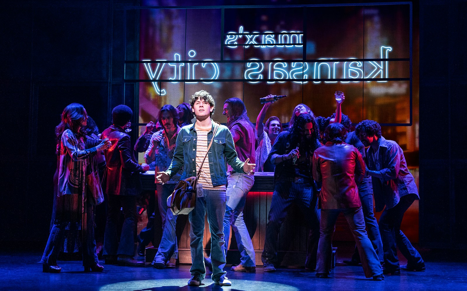 Young man on stage with a group in a bar setting, neon Kansas City sign in background, Almost Famous musical.