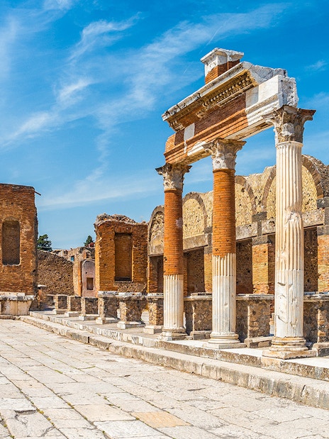 Ancient ruins of Pompeii with columns and archway, Mount Vesuvius in background.