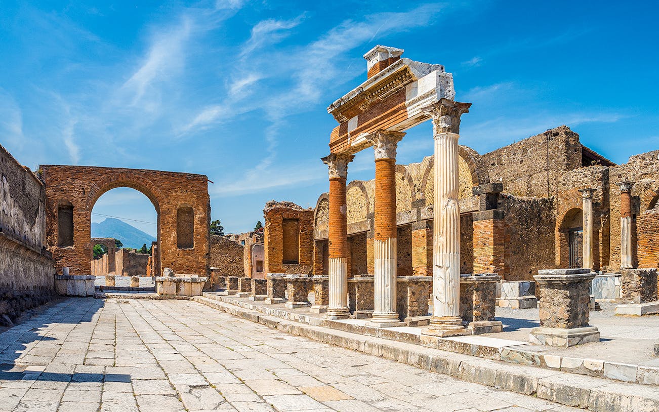 Ancient ruins of Pompeii with columns and archway, Mount Vesuvius in background.