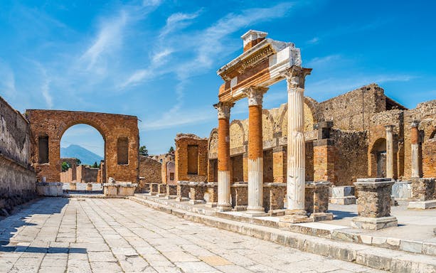 Ancient ruins of Pompeii with columns and archway, Mount Vesuvius in background.