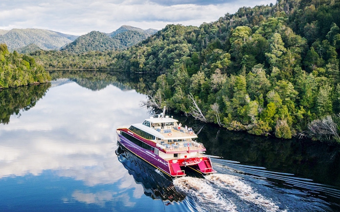 Red cruise boat on Gordon River, surrounded by lush forest and mountains, World Heritage site.