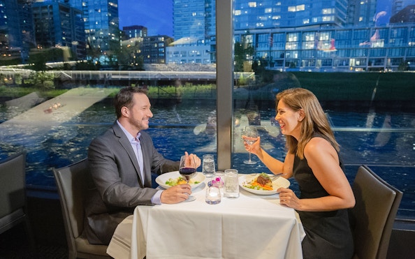 Couple dining by a window with city and water view at night.
