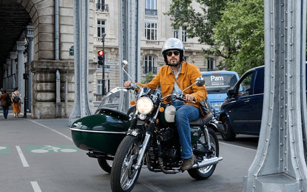 Man riding a motorcycle with sidecar under Paris bridge on a retro tour.