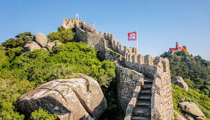 Moorish Castle ruins with steps and flags, Pena Palace in background, Sintra, Portugal.