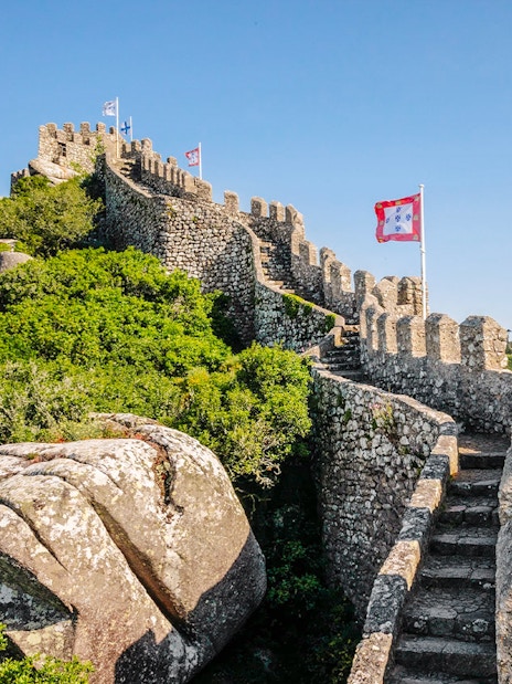 Moorish Castle ruins with steps and flags, Pena Palace in background, Sintra, Portugal.