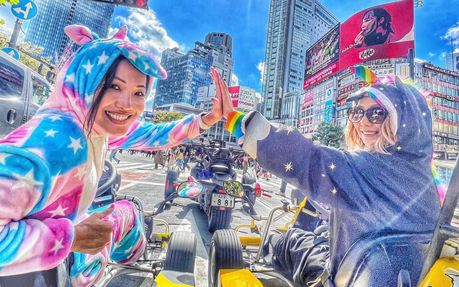 Two women in unicorn onesies high-five in go-karts on a Shibuya street.