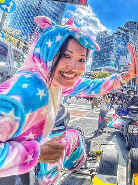 Two women in unicorn onesies high-five in go-karts on a Shibuya street.