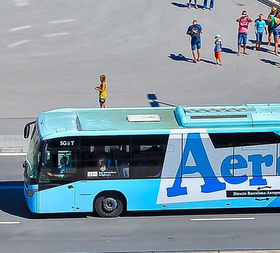 Aerobus driving on a city street with people waiting nearby.