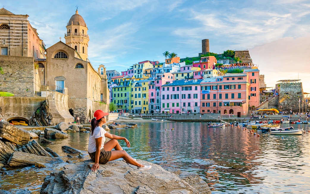 Tourist sitting on rocks overlooking colorful buildings in Vernazza, Cinque Terre.