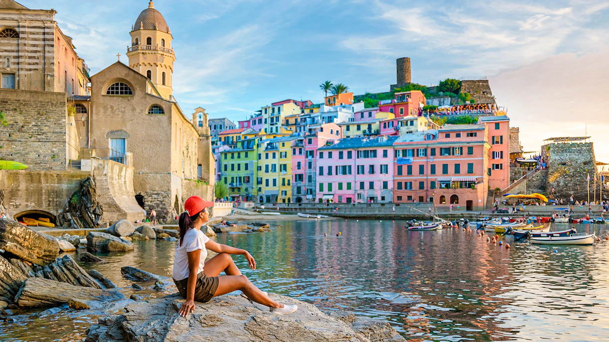 Tourist sitting on rocks overlooking colorful buildings in Vernazza, Cinque Terre.