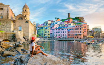 Tourist sitting on rocks overlooking colorful buildings in Vernazza, Cinque Terre.
