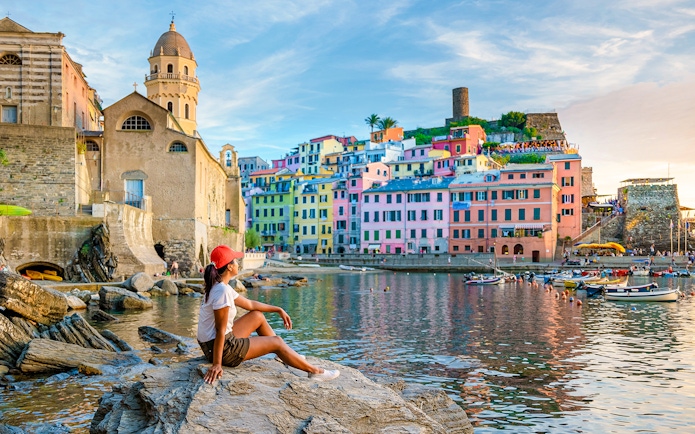 Tourist sitting on rocks overlooking colorful buildings in Vernazza, Cinque Terre.