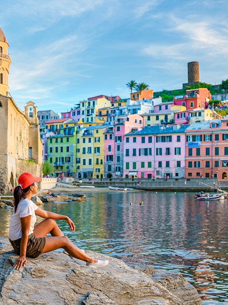 Tourist sitting on rocks overlooking colorful buildings in Vernazza, Cinque Terre.