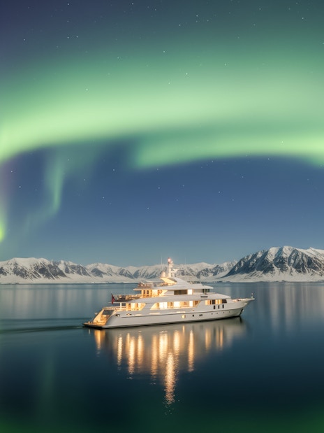 Yacht cruising under Northern Lights with snowy mountains in the background.