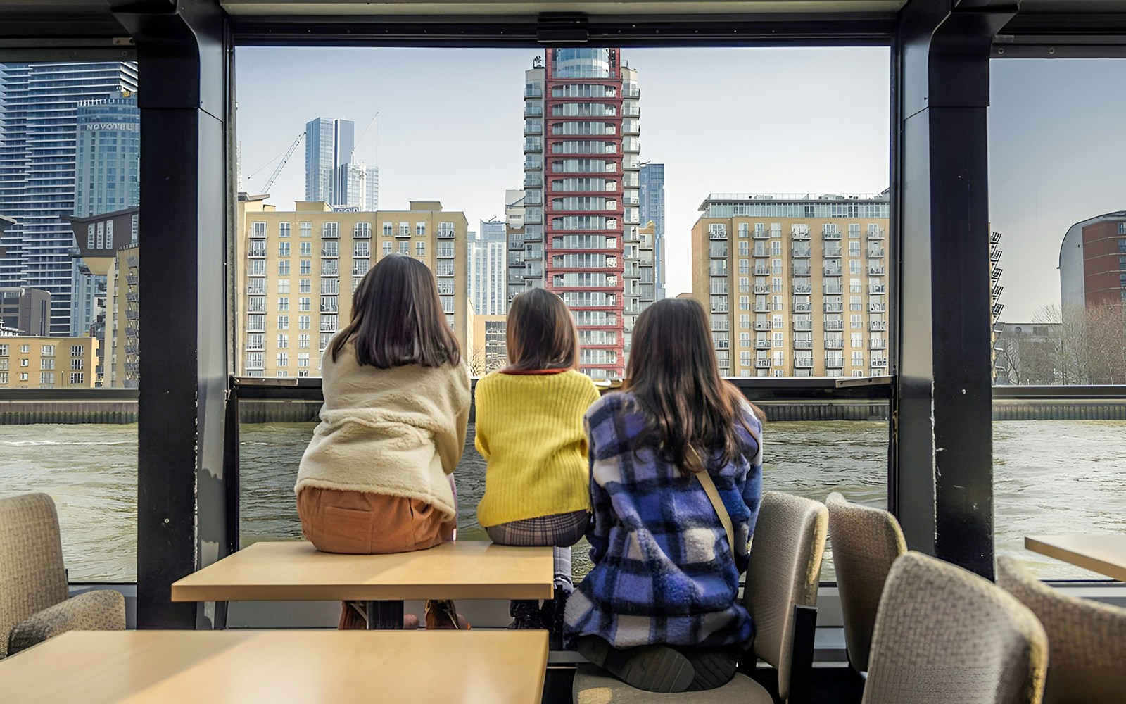 Three people seated inside a Thames River cruise boat, viewing modern London buildings.