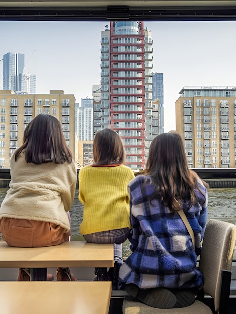 Three people seated inside a Thames River cruise boat, viewing modern London buildings.