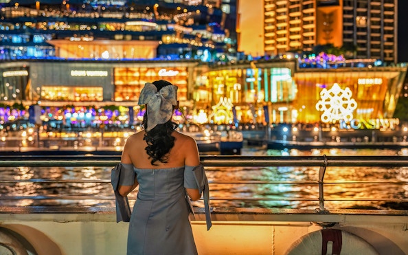 Lady viewing illuminated Bangkok buildings from Chao Phraya river cruise.